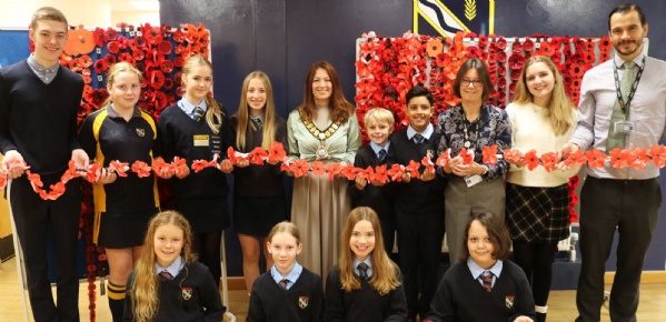 Group of school pupils and teachers holding a chain of red paper poppies in front of poppy displays on the wall, commemorating Remembrance Day.